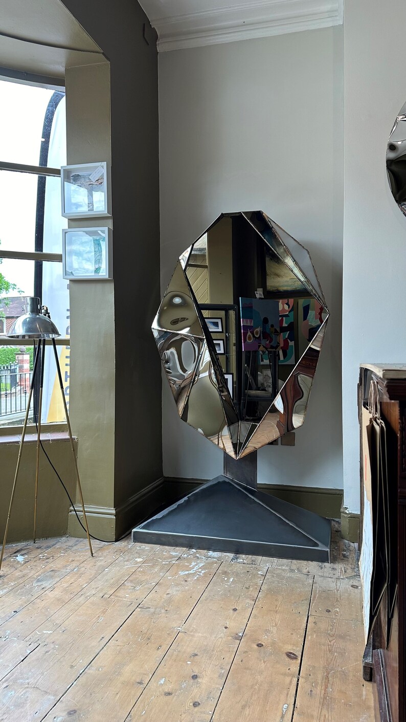 May include: A large, faceted, silver mirror with a geometric design. The mirror is standing on a black base in a room with wooden floors.