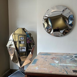 May include: Two silver-toned, abstract, geometric mirrors hang on a white wall. The mirror on the left is a faceted, diamond-shaped mirror. The mirror on the right is a round, wavy mirror. A dark wood cabinet with a drawer and two doors sits in front of the mirrors.