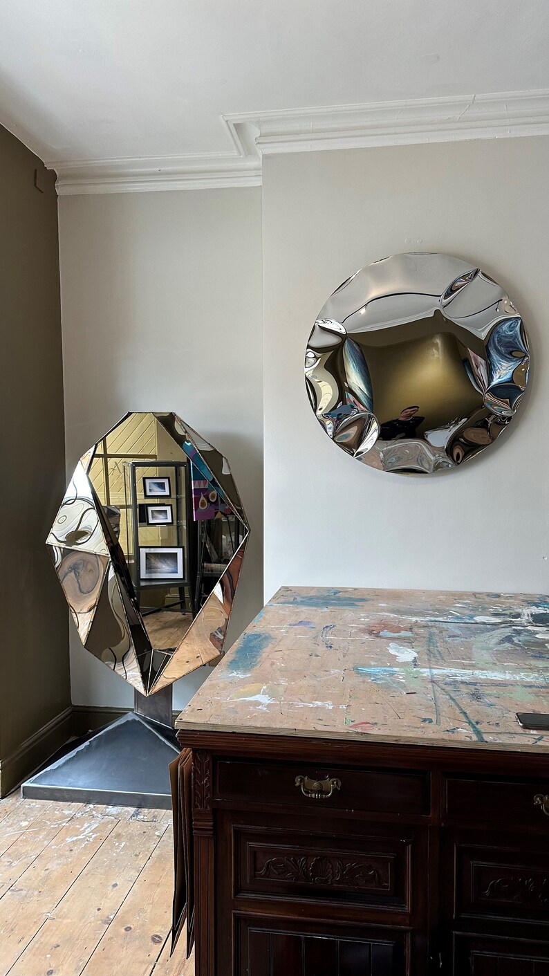 May include: Two silver-toned, abstract, geometric mirrors hang on a white wall. The mirror on the left is a faceted, diamond-shaped mirror. The mirror on the right is a round, wavy mirror. A dark wood cabinet with a drawer and two doors sits in front of the mirrors.