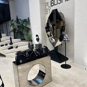 May include: A display of black handbags and shoes in a shop with a large, geometric, silver mirror with the text "PUBLISHED BY" on the wall behind it.
