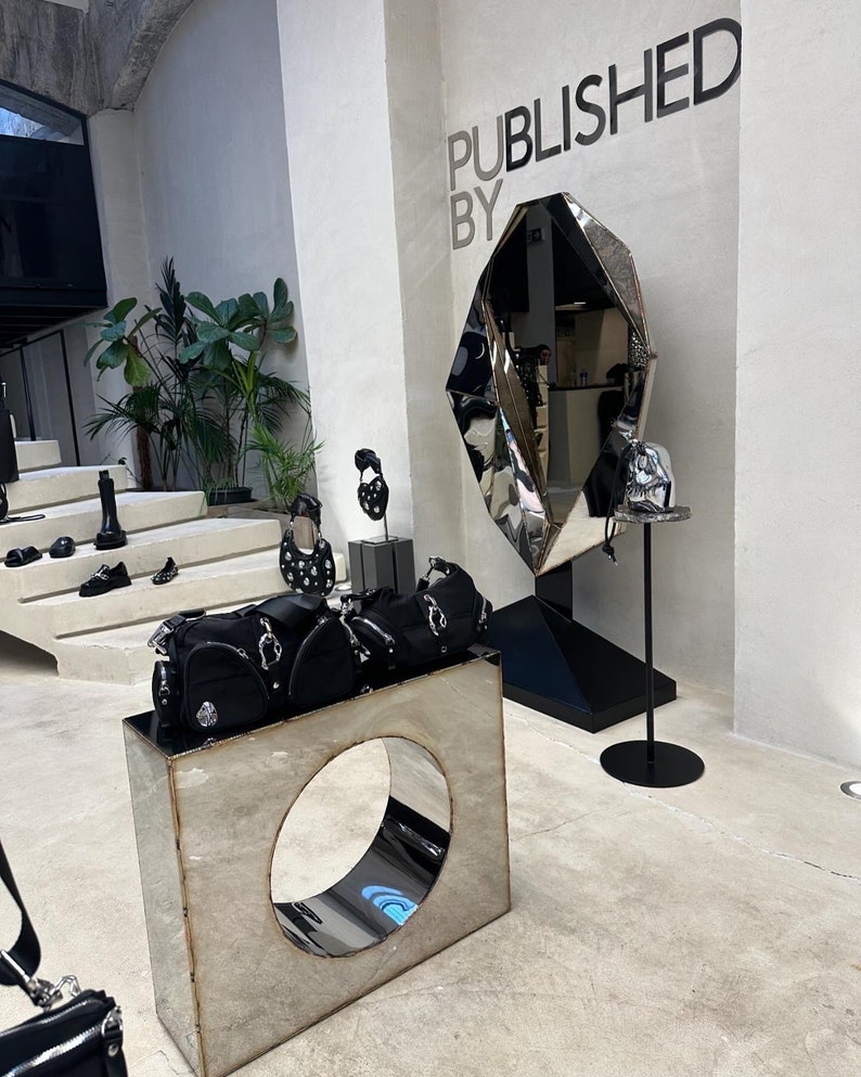 May include: A display of black handbags and shoes in a shop with a large, geometric, silver mirror with the text "PUBLISHED BY" on the wall behind it.
