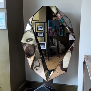 May include: A large, faceted, silver mirror with a geometric shape, standing on a black metal base. The mirror is reflecting the interior of a room with artwork on the walls.
