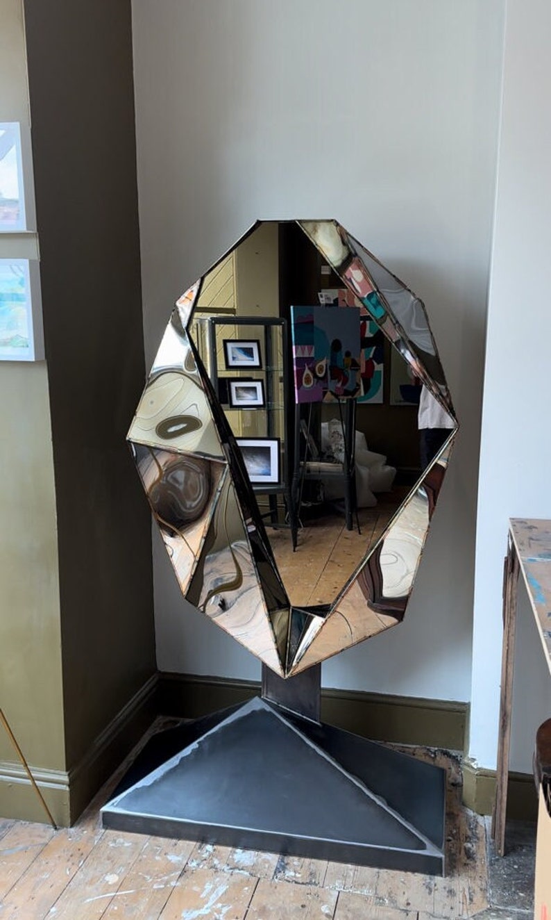 May include: A large, faceted, silver mirror with a geometric shape, standing on a black metal base. The mirror is reflecting the interior of a room with artwork on the walls.