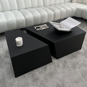 May include: Two black triangular coffee tables with a white candle on one table and a book on the other table.