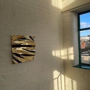 Könnte beinhalten: Ein zerknittertes Metallblatt in Goldfarbe ist an einer weißen Backsteinmauer befestigt. Die Wand befindet sich in einer Ecke mit einem Fenster auf der rechten Seite. Das Fenster hat einen grünen Rahmen und einen Blick auf Dächer und einen blauen Himmel.