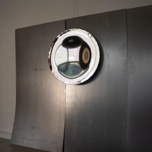 May include: A circular, highly reflective convex mirror with a polished silver finish, suspended against a large, dark grey, rectangular metal panel. The mirror surface displays distorted reflections of the surrounding environment in shades of black.