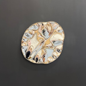 Könnte beinhalten: Ein metallisches, unregelmäßig geformtes Dekorationsobjekt mit einer reflektierenden, gewellten Oberfläche. Das Objekt hat einen goldfarbenen Rand und reflektiert die Umgebung, wodurch ein verzerrtes, abstraktes Muster entsteht. Der Hintergrund ist einfarbig dunkelgrau.