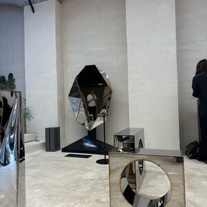 May include: A large, faceted, silver-coloured, geometric sculpture with a reflective surface. The sculpture is in a room with white walls and a concrete ceiling. There are two smaller, silver-coloured, geometric sculptures with circular openings in the room.