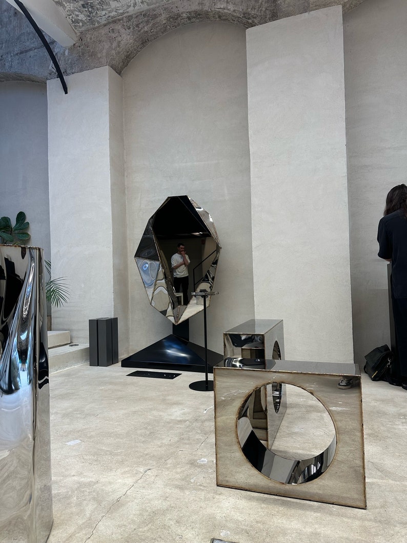May include: A large, faceted, silver-coloured, geometric sculpture with a reflective surface. The sculpture is in a room with white walls and a concrete ceiling. There are two smaller, silver-coloured, geometric sculptures with circular openings in the room.