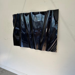 Könnte beinhalten: Eine schwarze, abstrakte Wandskulptur mit einer gewellten, wellenförmigen und strukturierten Oberfläche. Die Skulptur hängt an einer weißen Wand.