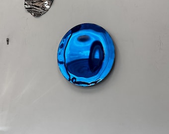 May include: Two abstract wall art pieces. A large, round, reflective blue disc and a smaller, irregularly shaped, silver metal piece are mounted on a white wall. The blue disc has a glossy, liquid-like appearance, reflecting the surrounding environment.