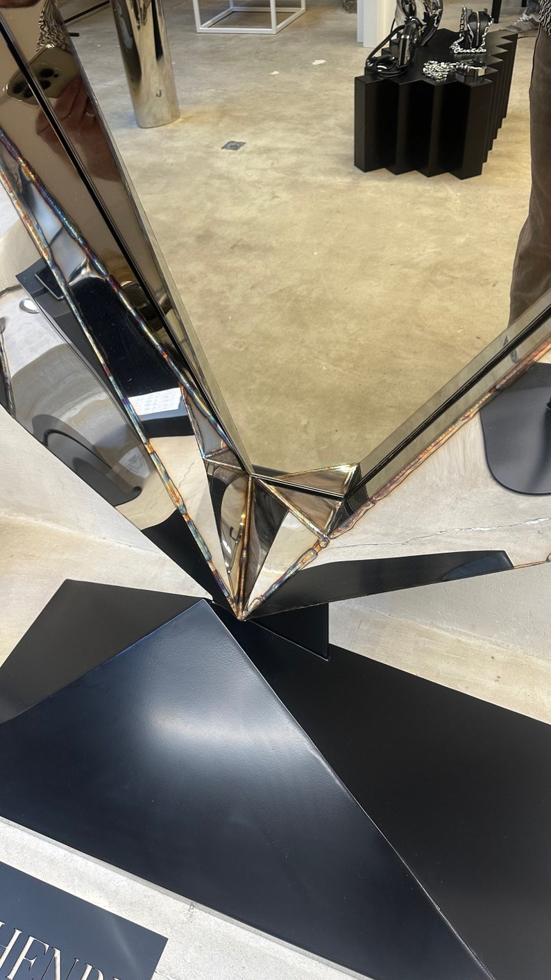 May include: A large, silver, geometric mirror with a black base. The mirror is made of multiple pieces of metal that are joined together to create a unique and modern design.