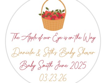 May include: A round sticker with a basket of red apples and green leaves. The text "The Apple of our Eye is on the Way" is in a cursive font. Below it, "Daniela & Seth's Baby Shower" and "Baby Smith June 2025" are written. The date "03.23.26" is at the bottom.