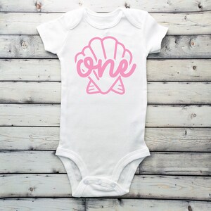 May include: White baby onesie with a pink seashell design and the word "one" inside the shell.