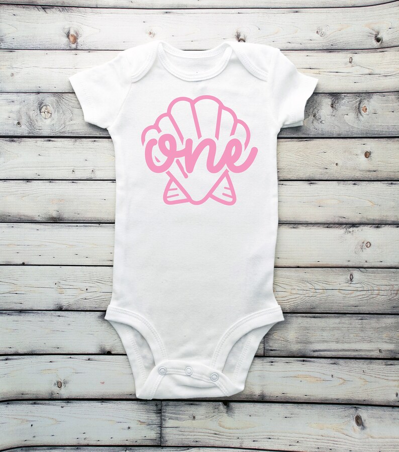 May include: White baby onesie with a pink seashell design and the word "one" inside the shell.