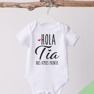 May include: White baby onesie with black text that reads "Hola Tia Nos Vemos Pronto" with a small red heart before "Hola".