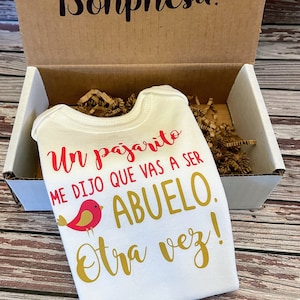 May include: A white onesie with a red bird and gold text that reads "Un pajarito me dijo que vas a ser ABUELO otra vez!" The onesie is in a brown cardboard box with shredded paper.