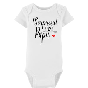 May include: White baby onesie with short sleeves and a black graphic that says "!Sorpresa! Serás Papa" with a red heart and arrow.