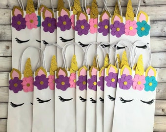 May include: A set of 15 white paper gift bags decorated with unicorn faces. Each bag has a gold glitter horn, pink and purple flowers, and black eyelashes. The bags have white string handles.