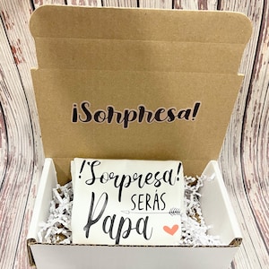 May include: A white box with a white card inside. The card has black text that says "¡Sorpresa! Serás Papa" with a pink heart and an arrow. The box is open and the card is visible. The box is sitting on a wooden surface.