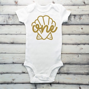 May include: White baby onesie with a gold seashell design and the word "one" inside the shell.