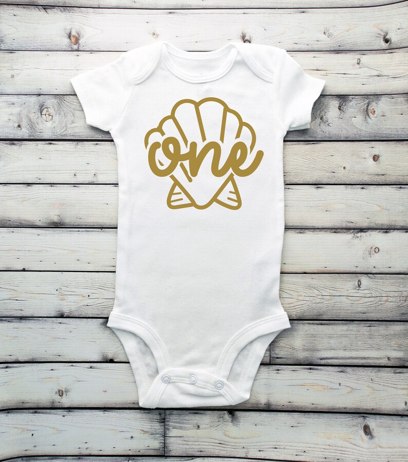 May include: White baby onesie with a gold seashell design and the word "one" inside the shell.