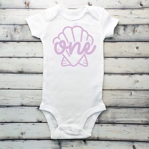 May include: White baby onesie with a purple seashell design and the word "one" inside the shell.
