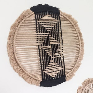 May include: A round wall hanging made of natural fibre rope with a black geometric pattern woven into the centre. The rope has a fringe around the edge.