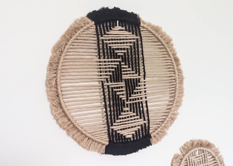 May include: A round wall hanging made of natural fibre rope with a black geometric pattern woven into the centre. The rope has a fringe around the edge.
