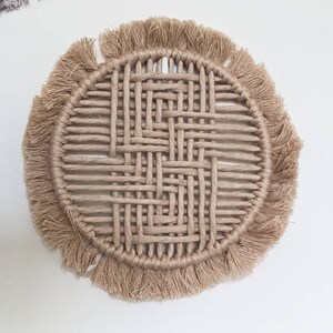 May include: A round woven coaster with a fringe border. The coaster is made of natural fibres and has a light brown colour.