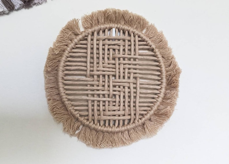 May include: A round woven coaster with a fringe border. The coaster is made of natural fibres and has a light brown colour.