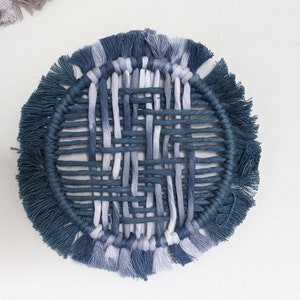 May include: A round woven wall hanging with a blue and white colour scheme. The hanging has a fringe border and a woven centre.