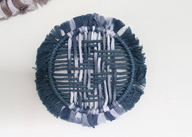 May include: A round woven wall hanging with a blue and white colour scheme. The hanging has a fringe border and a woven centre.