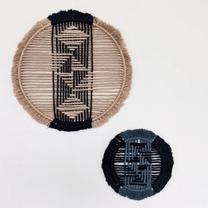May include: Two circular wall hangings made of woven macrame. The larger one is beige with a black geometric pattern. The smaller one is black with a blue geometric pattern.