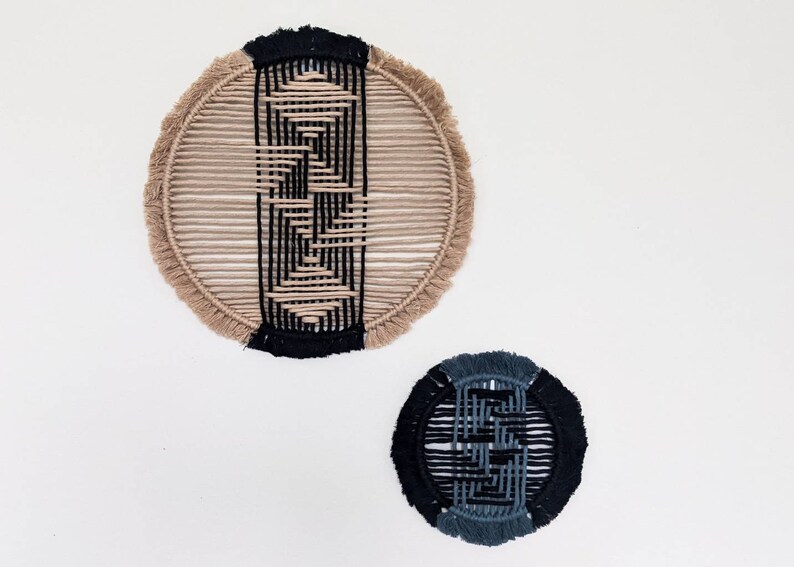 May include: Two circular wall hangings made of woven macrame. The larger one is beige with a black geometric pattern. The smaller one is black with a blue geometric pattern.