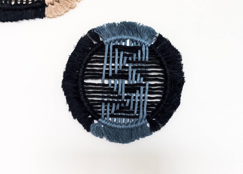 May include: A round, woven wall hanging with a black and blue geometric pattern. The hanging has a fringe border.