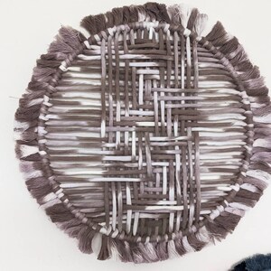 May include: A round woven wall hanging with a fringe border. The hanging is made of white and brown yarn and has a geometric pattern.