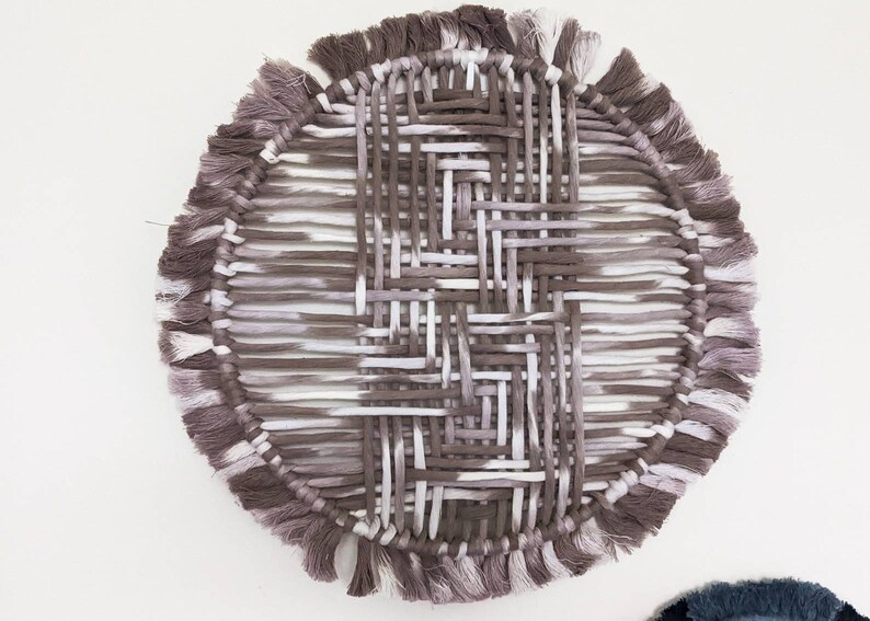 May include: A round woven wall hanging with a fringe border. The hanging is made of white and brown yarn and has a geometric pattern.
