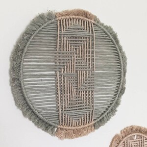 May include: A round wall hanging made of woven macrame with a geometric pattern. The macrame is in shades of light green and beige with a fringe border.