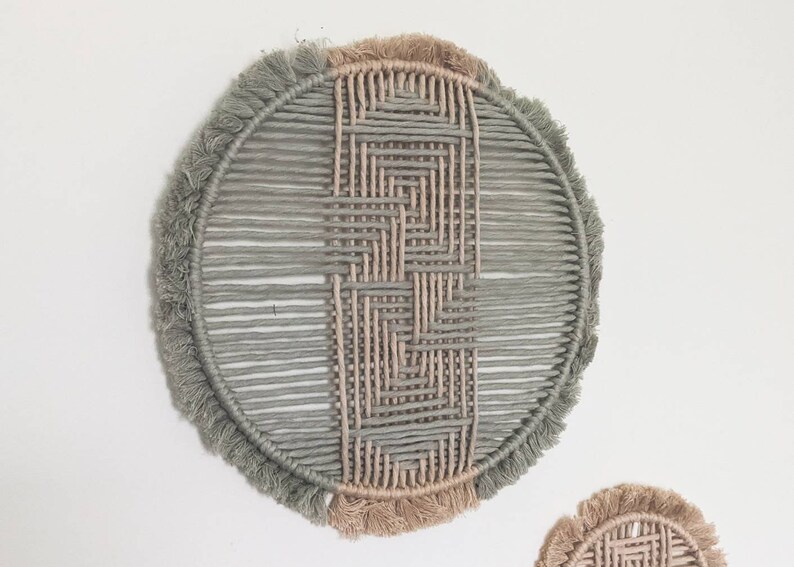 May include: A round wall hanging made of woven macrame with a geometric pattern. The macrame is in shades of light green and beige with a fringe border.