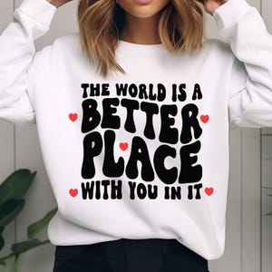 May include: White sweatshirt with a black graphic that says "The world is a better place with you in it". The graphic is in a retro font with red hearts on either side.
