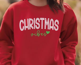 May include: A red sweatshirt with the words "CHRISTMAS vibes" in white and green. The word "CHRISTMAS" is in a dotted font, and the word "vibes" is in a script font with a green heart.