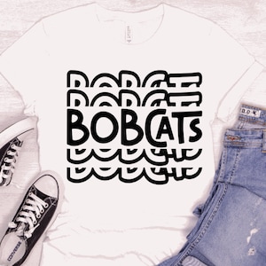 May include: White t-shirt with a black graphic design that says "BOBCATS" in a repeating pattern.