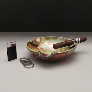 May include: A ceramic ashtray with a green, yellow, and orange glaze. A cigar is resting in the ashtray. A silver cigar cutter and a black lighter are also pictured.