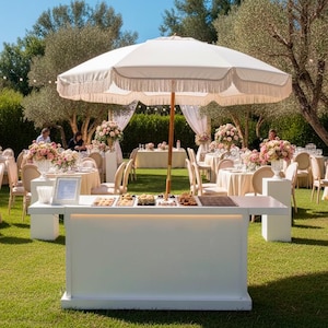 May include: An outdoor event setup with tables, chairs, and a white dessert bar. A large white umbrella with fringe provides shade. Tables are decorated with floral arrangements. The scene is set on a green lawn under a blue sky.
