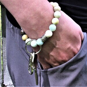 May include: A beaded bracelet with a silver charm and a key chain attached. The bracelet is made of light blue, white, and brown beads.