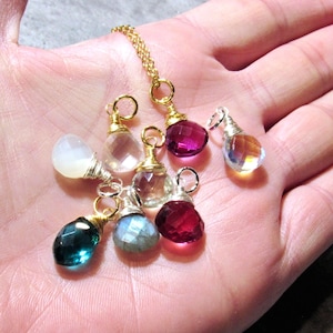 May include: A collection of gemstone pendants in various colors, including clear, pink, green, and blue, with gold-colored metal accents. The pendants are teardrop-shaped and are displayed on a gold chain. The gemstones are faceted, reflecting light.