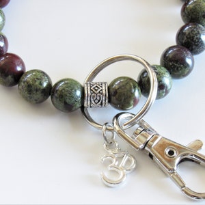 May include: A green and brown beaded keychain with a silver key ring, a silver charm with an Om symbol, and a silver clasp.
