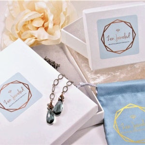 May include: A white box with a blue and gold logo that says "Zen Jewelled" and the text "Beautiful Jewelry for Health & Well-being". The box contains a silver chain necklace with a teardrop-shaped blue gemstone pendant. The necklace is packaged in a white box with a blue velvet pouch.
