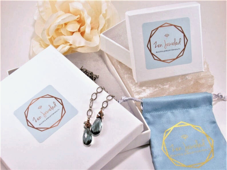 May include: A white box with a blue and gold logo that says "Zen Jewelled" and the text "Beautiful Jewelry for Health & Well-being". The box contains a silver chain necklace with a teardrop-shaped blue gemstone pendant. The necklace is packaged in a white box with a blue velvet pouch.